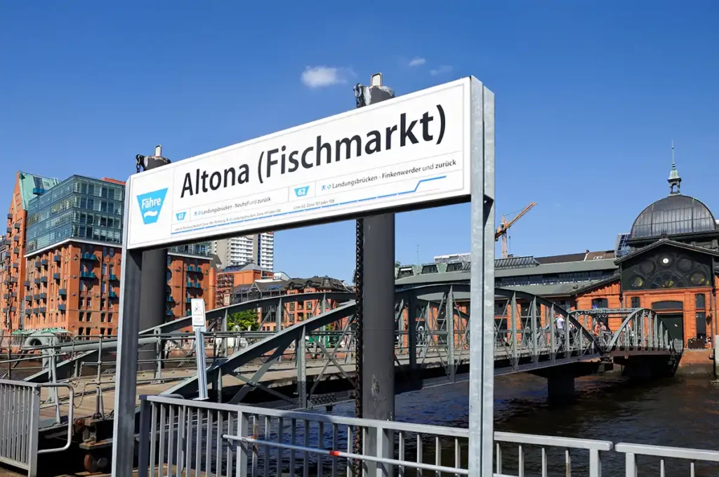 A ferry stop in Hamburg. The sign reads Altona Fischmarkt and there is a large brick building with a dome in the background.