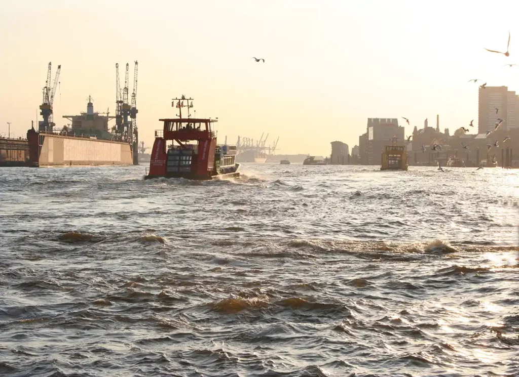 Hamburg ferries on the River Elbe. A small boat is on a wide river, with dock cranes on one bank and buildings on the other.