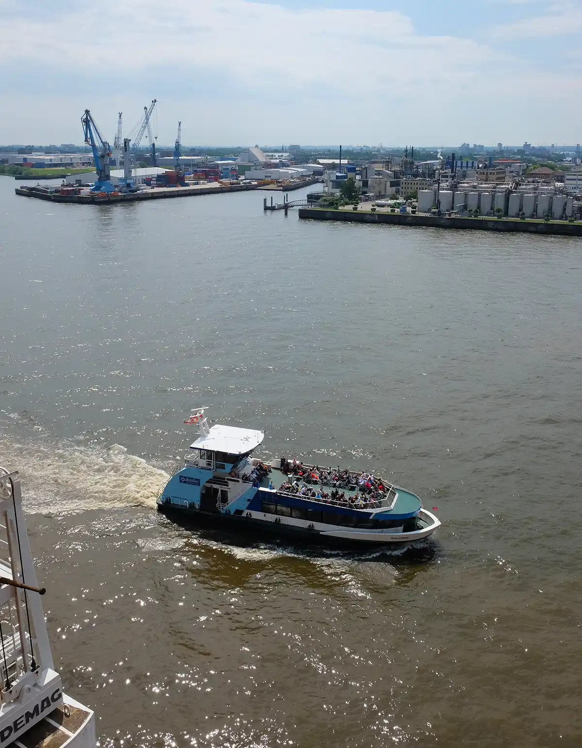 A Hamburg ferry leaving the Elbphilharmonie stop. The ferry is seen from above. It has an open top deck full of people. There are cranes and docks in the background. 