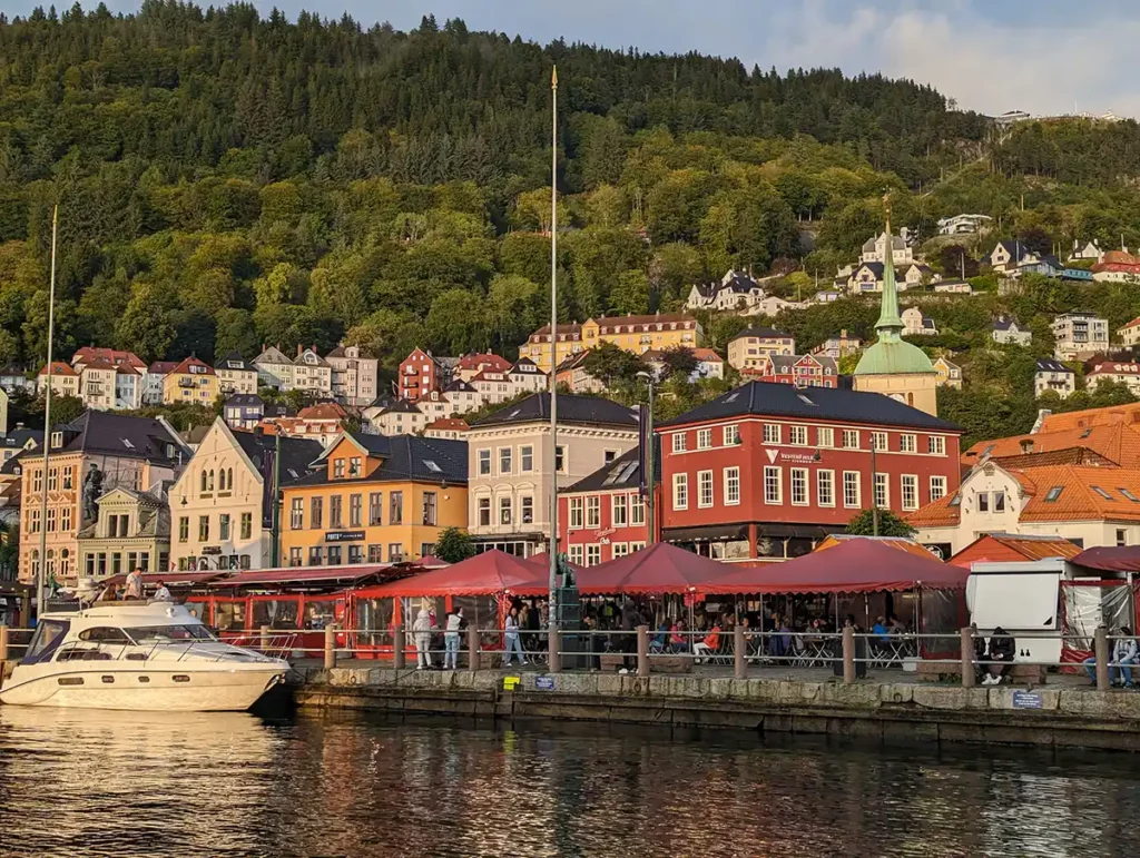 The harbour in Bergen. Colourful, low-rise buildings in red, yellow and white are clustered around a harbour. There is a thickly wooded hill, with more colourful buildings leading up the hill. 