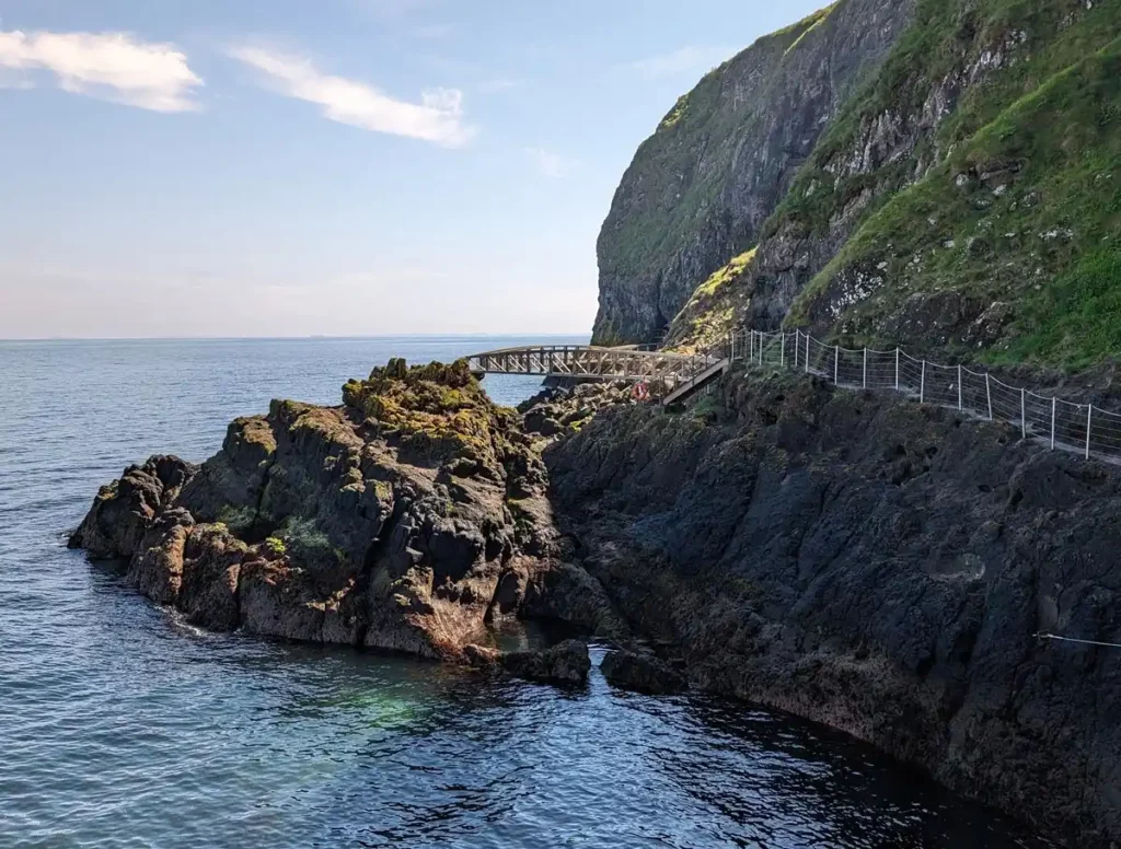 A rocky cliff with the sea below. There's a path running along the cliff edge, a few metres above the waterline. The path then leads onto two bridges which stick out above the water.