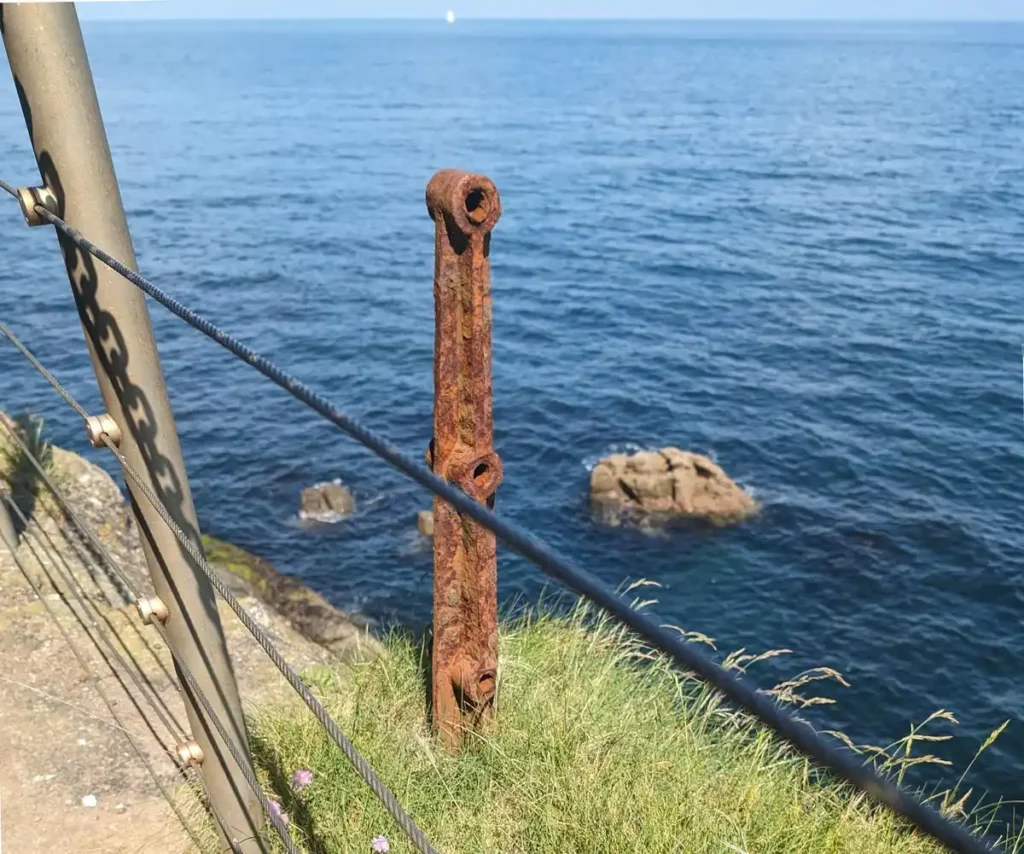 An old, rusty post on a cliff above the sea. A modern equivalent is next to it but much taller. 