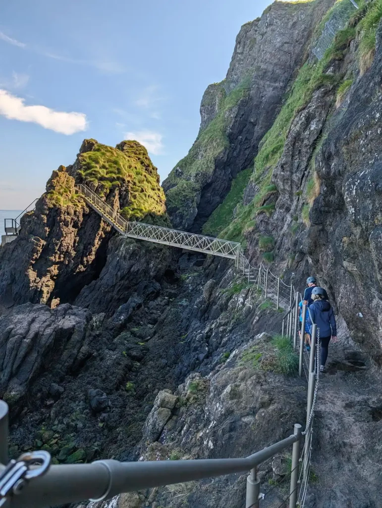 A narrow rocky path along the base of a sea cliff, leading to a metal bridge.