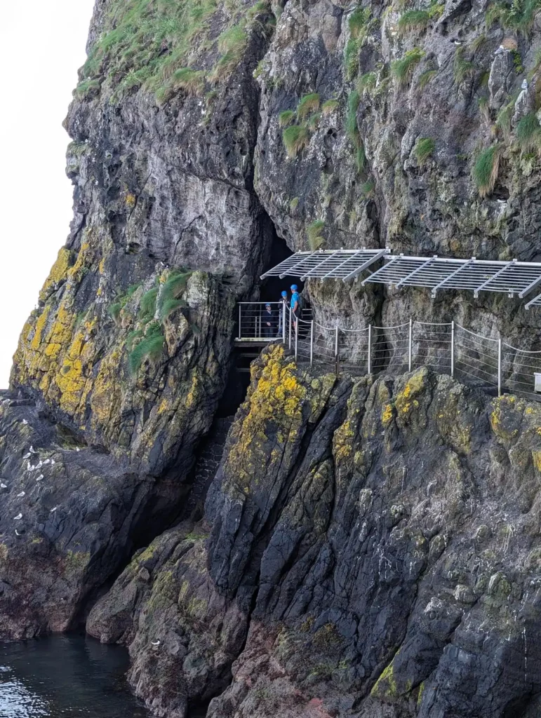 A rocky cliff face above the sea. Some people are emerging from a crack in the rock onto a narrow path around the cliff.