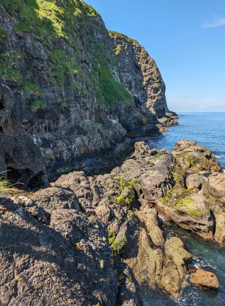 A narrow path snaking around the base of a sea cliff, with a rocky outcrop in front. 