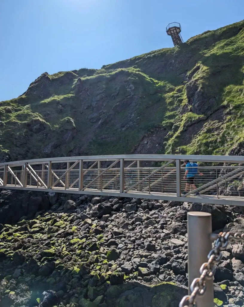 A man walking across a metal bridge at the base of a cliff. There's a metal viewing platform jutting out into the air at the top of the cliff.