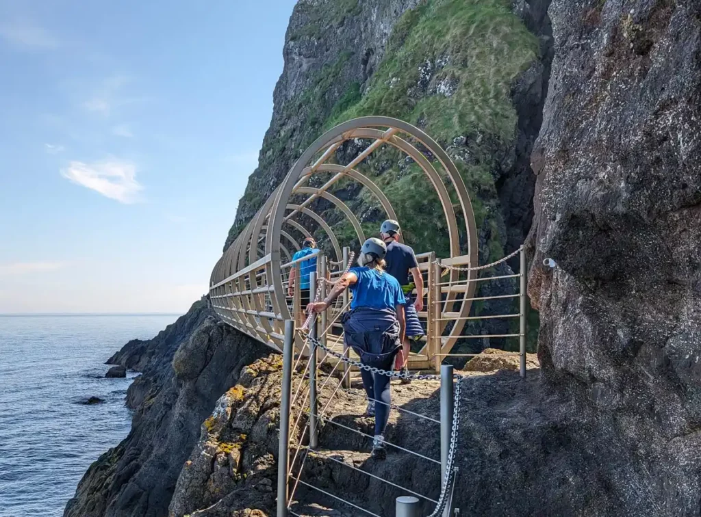 The Tubular Bridge is one of the highlights of the Gobbins cliff path walk. A tubular metal bridge at the base of cliffs. Some people in hard hats are walking across the bridge.