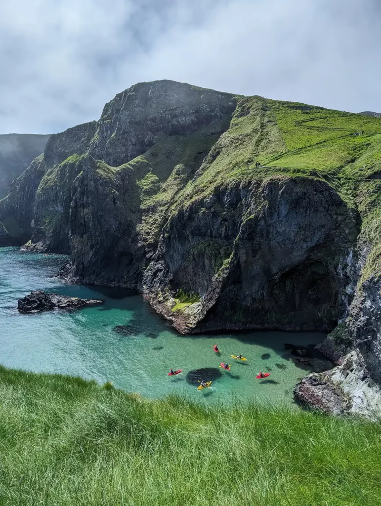 Green cliffs and a turquoise sea. There are a group of people in kayaks at the bottom of the cliffs.