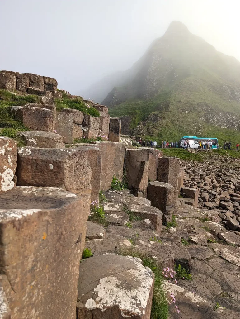 Basalt columns at the Giant's Causeway. There is a bus in the background and a spiky green cliff, which is partially shrouded in mist. 