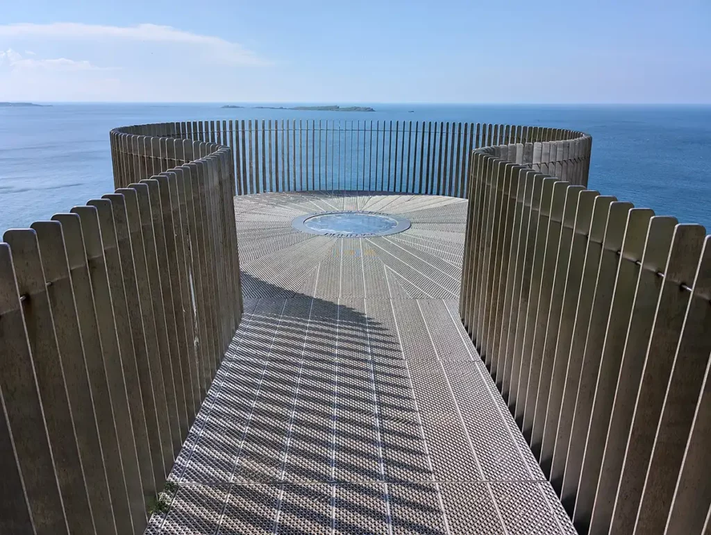 A metal walkway with a circular viewing platform at the end, looking out over the sea