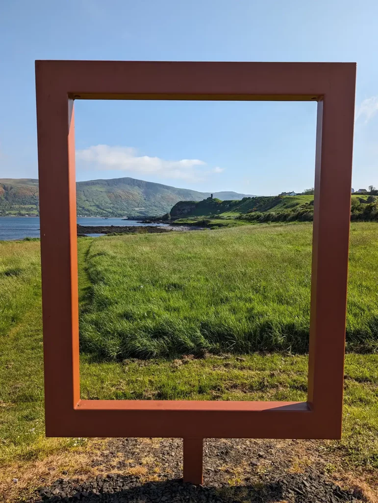 A large, rusty red metal frame, framing a view of a ruined castle on a hill, green mountains and the sea