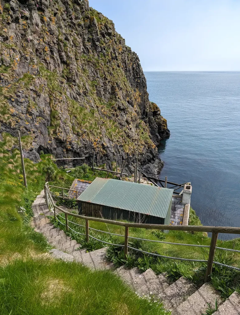 An old cottage at the bottom of a steep sea cliff. A flight of steps leads to the cottage.