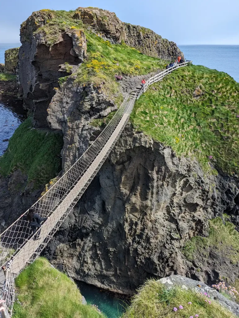 A narrow rope bridge with a person crossing, suspended between two rocky cliffs with the sea below