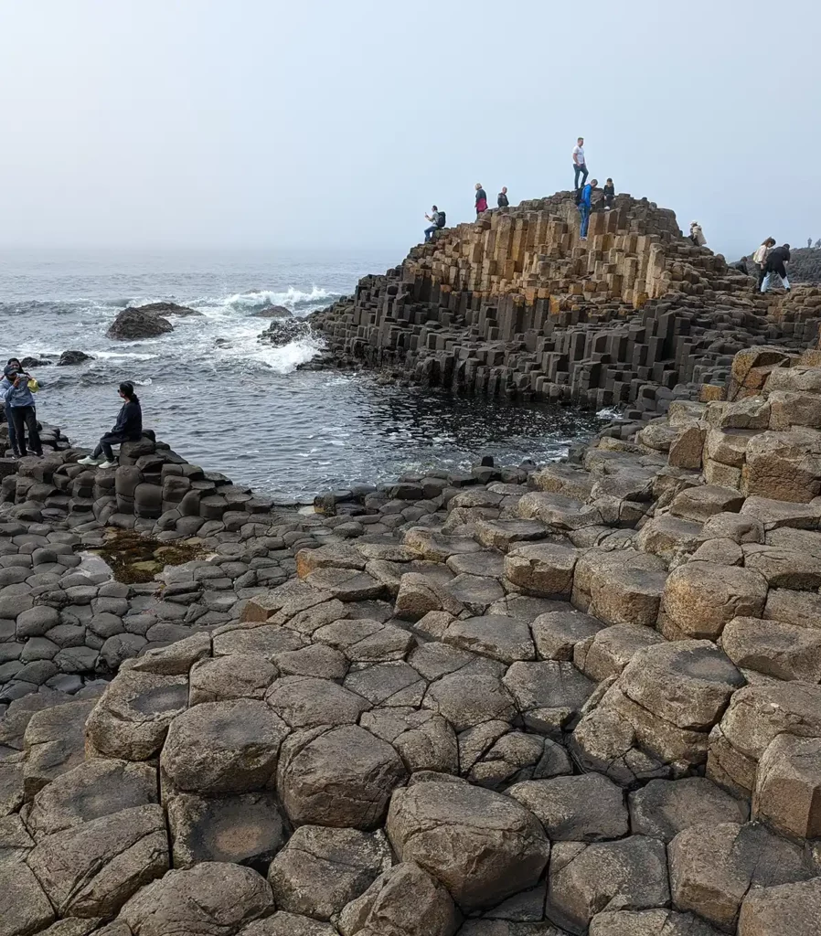 The hexagonal stones by the edge of the sea. There is a taller section in the background and people climbing on the rocks.