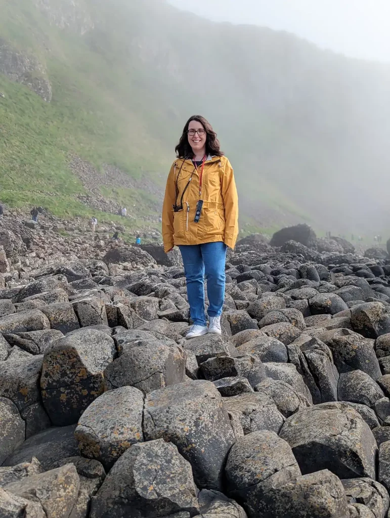 A woman (me!) wearing a bright yellow jacket and jeans standing on top of rocks at the Giant's Causeway, Northern Ireland