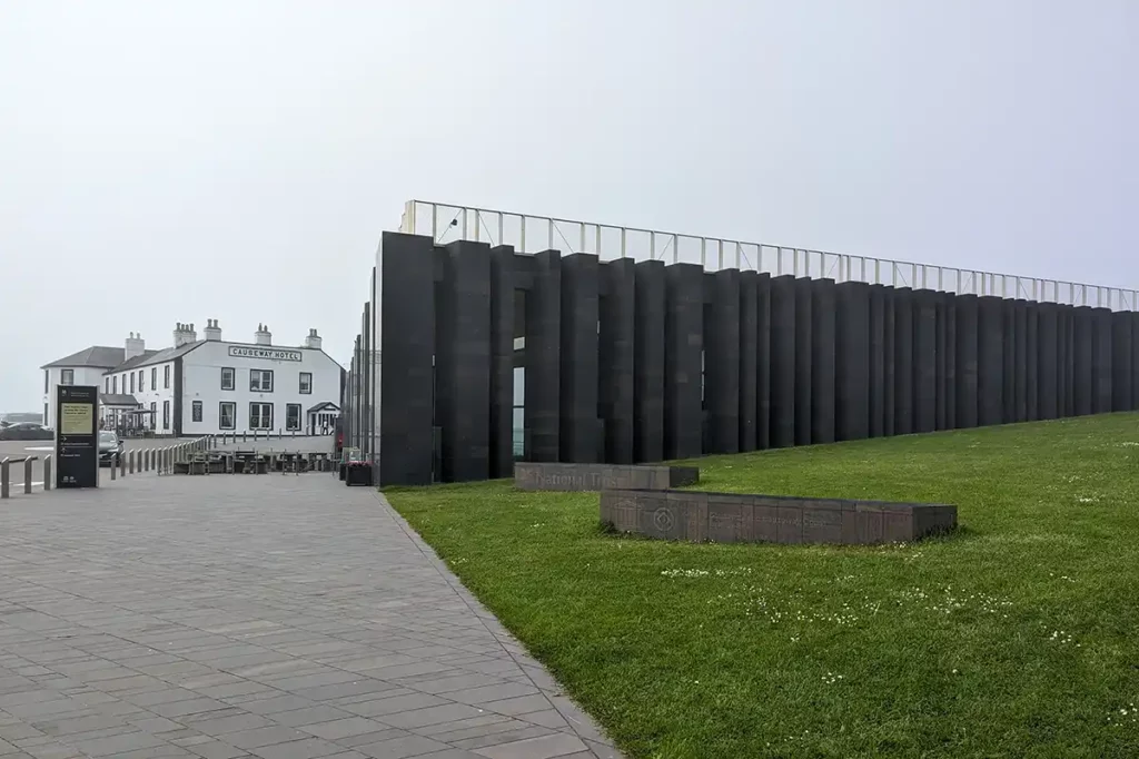 A large, modern, black building with a path and grass in front. An older hotel is in the background.