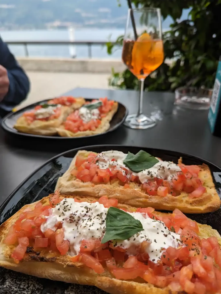 Enjoying aperitivo in Bellano by Lake Como. Two people are sat at a table eating bruschetta with tomatoes. There's an Aperol Spritz in the background.