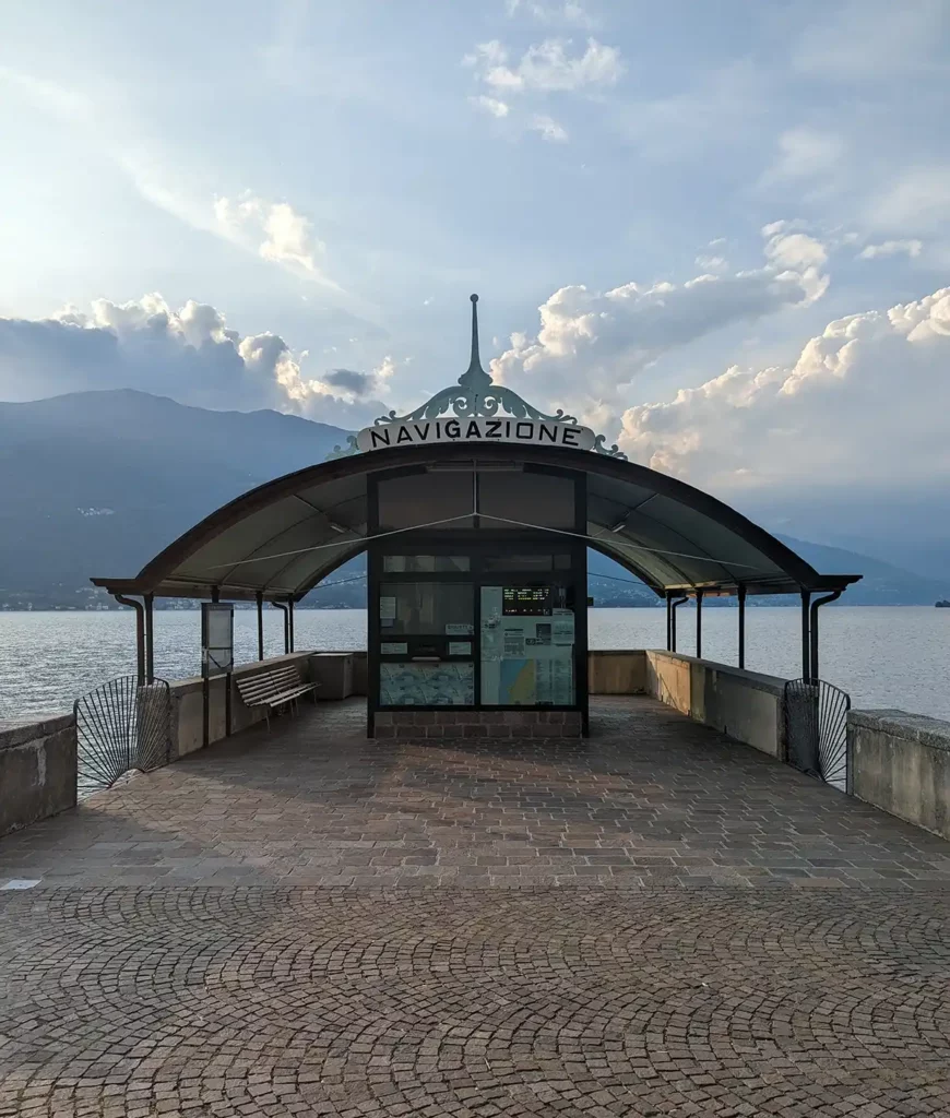 The ferry dock in Bellano. A pretty building with an ornate roof sits on a wooden jetty sticking out into a lake with mountains beyond.