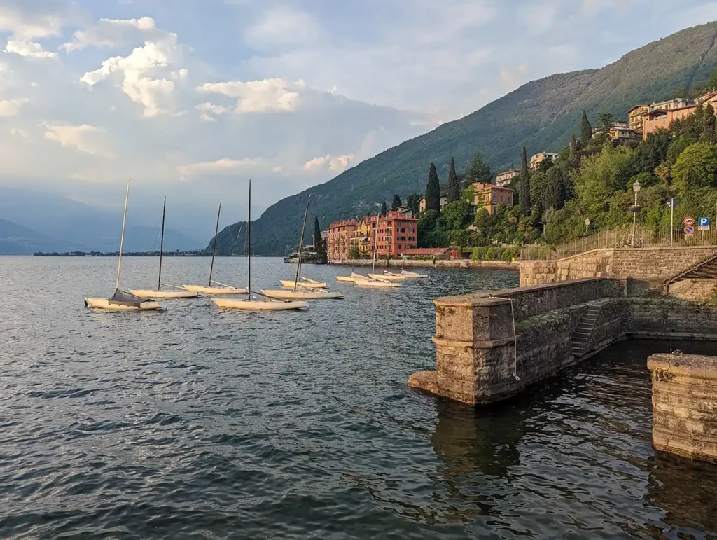 Golden hour in Bellano, a small village on Lake Como. The image shows a little harbour on the lake, with old-fashioned buildings in the distance and pine trees on the hills leading down to the lake. 