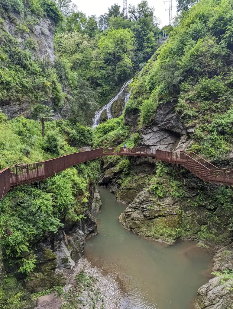 A deep natural, tree-lined gorge with a river in the bottom. There are walkways around the edges and a waterfall in the distance. 