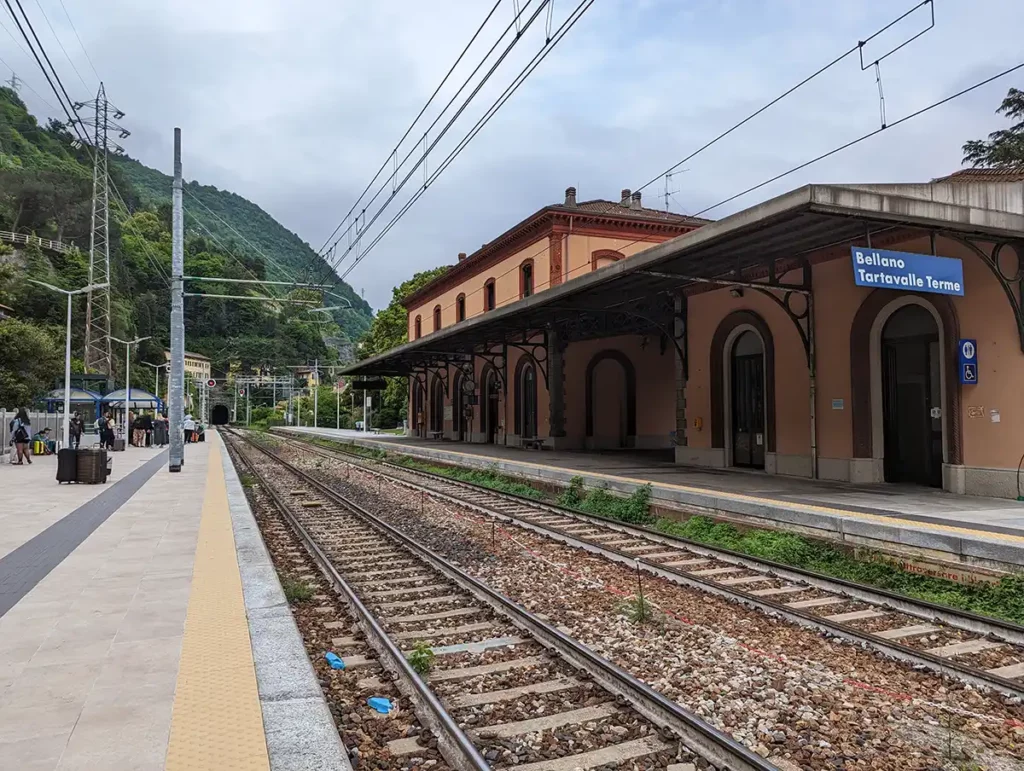 Bellano-Tartavalle Terme station. Two railway lines stretch out into the distance, leading to a tunnel in the hillside. A station building is on the opposite platform to where the photographer is standing.  