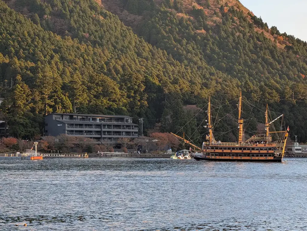 Rakuten Stay Fujimi Terrace from near the Hakone Shrine Torii gate. A dark-coloured apartment building sits between the lake and a forested hillside in Hakone, Japan. A pirate ship is crossing the lake in front of the apartment building. 