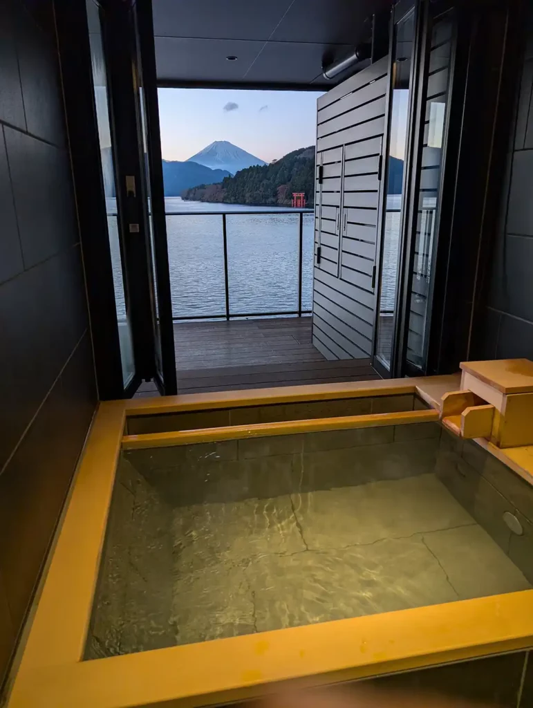 A wooden hot spring bath in a modern tiled bathroom. The balcony doors are open, and there is a view of a lake, a red Torii gate and Mount Fuji beyond.