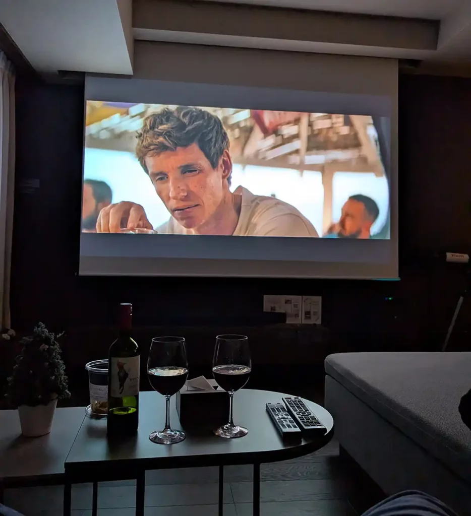 A projector screen playing a TV show in a dark living room. A bottle of wine and two glasses are on a coffee table in front of a sofa.