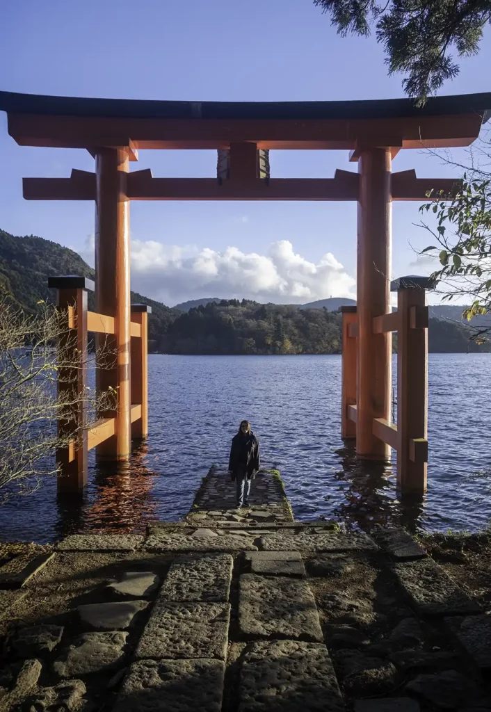 A woman stood underneath a read Torii gate on a lake, with forested hills in the background