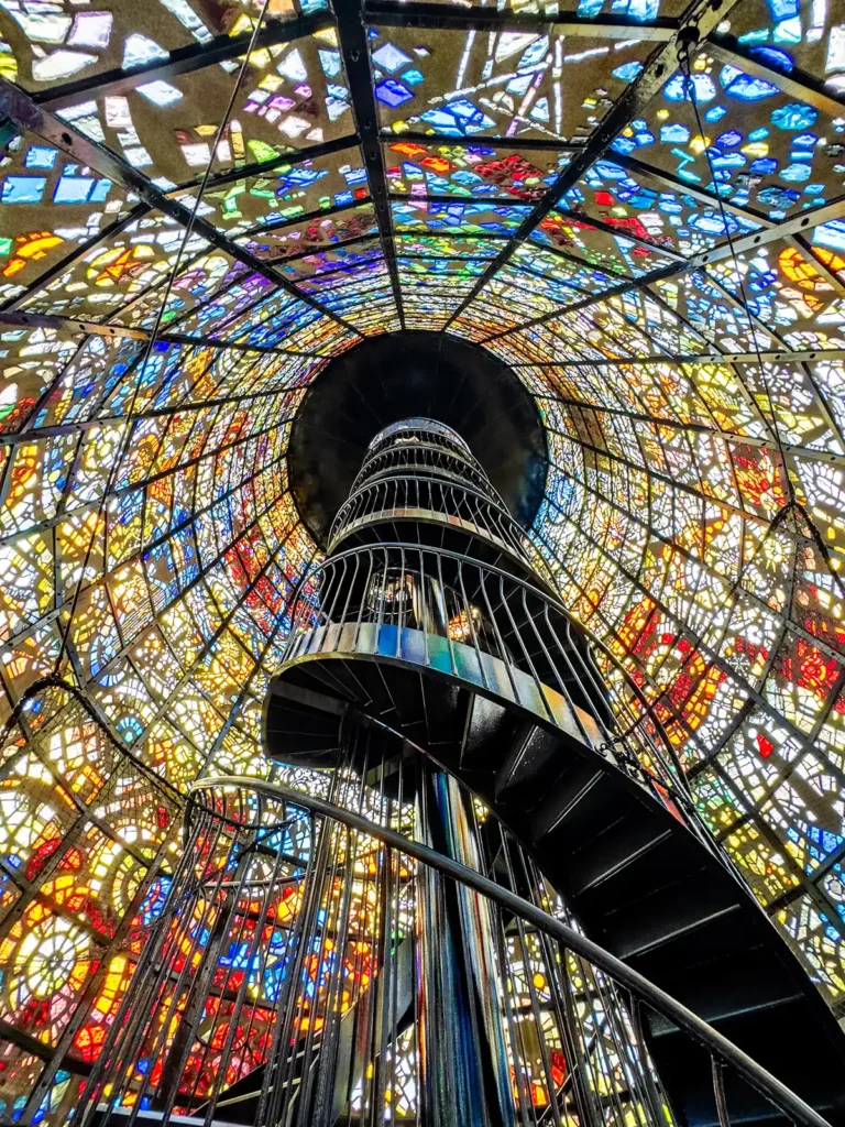 A shiny metal spiral staircase up a tower made of colourful stained glass