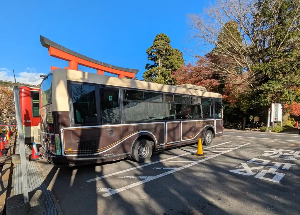 A brown bus parked up at a small bus station. There is a red torii gate crossing the road behind. 