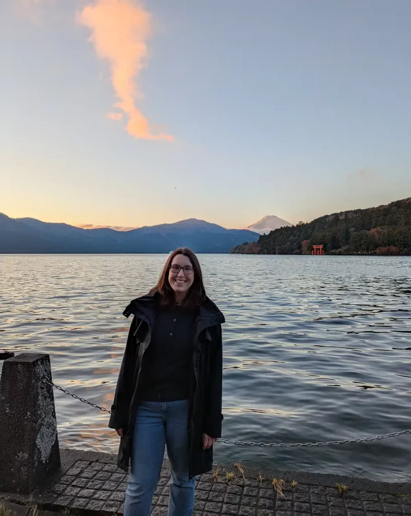 A woman stood in front of a lake, with a snow-capped triangular mountain in the background.