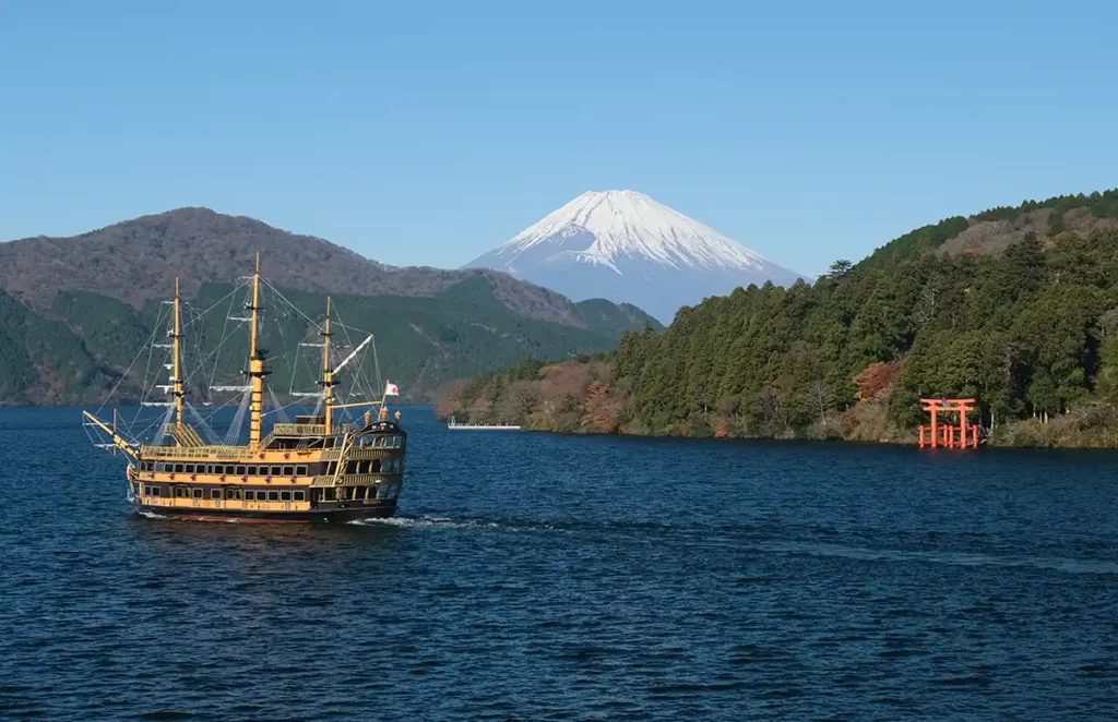 A pirate ship sailing across Lake Ashi in Hakone, Japan, with a red torii gate by the lake shore, forested mountains and a snow-capped Mount Fuji in the distance.