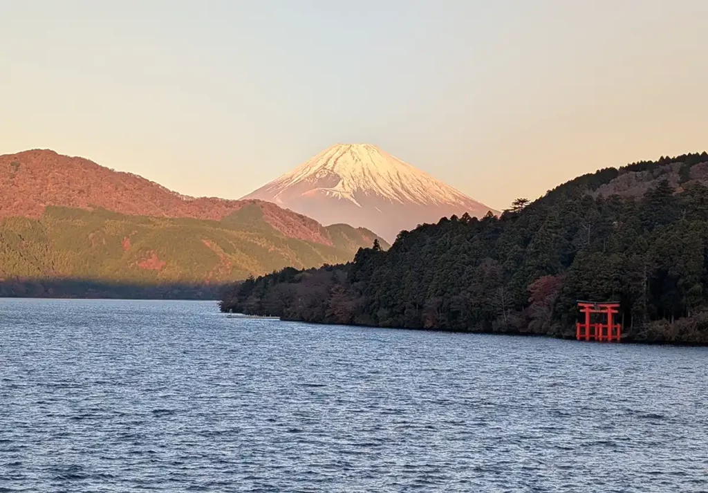 A view of Mount Fuji at sunrise - one of the benefits to staying overnight in Hakone vs taking a day trip from Tokyo. A huge, triangular, snow-capped mountain is bathed in pink light. In the foreground, there's a red shrine by the side of a lake.