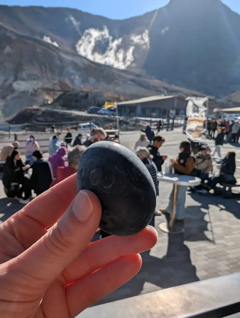 A woman's hand holding a black egg. There are lots of visitors in the background and the hillside beyond is steaming with volcanic sulphur vents.