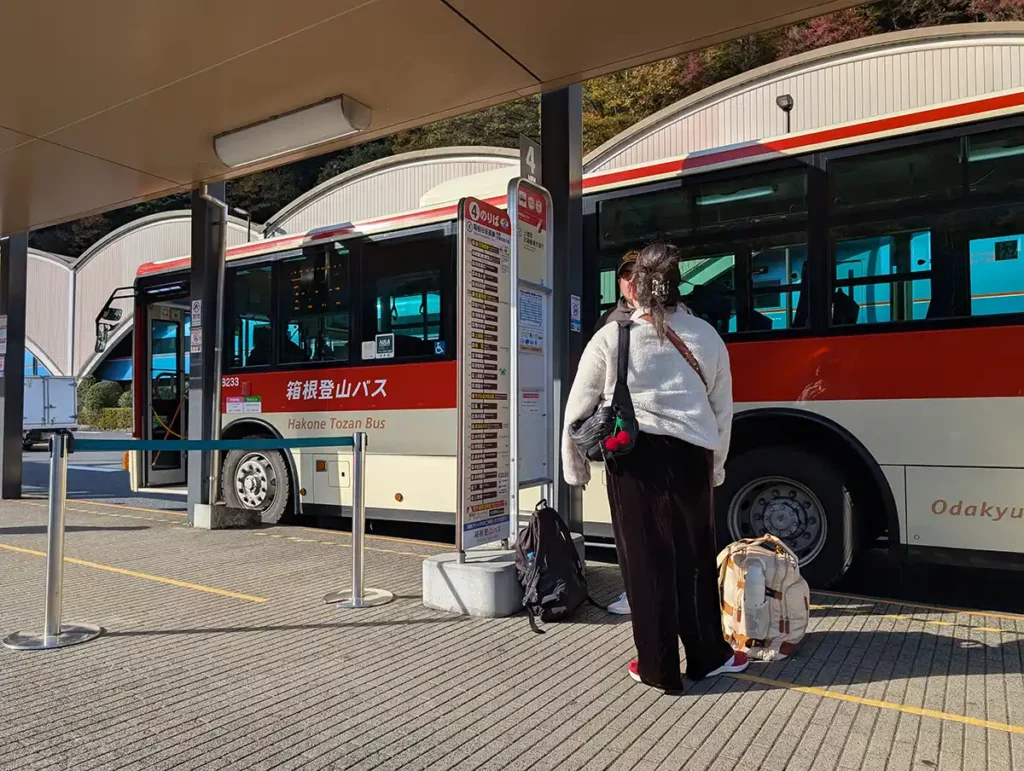 A bus stop in Hakone-Yumoto. There is a red and white bus parked up and people waiting. 