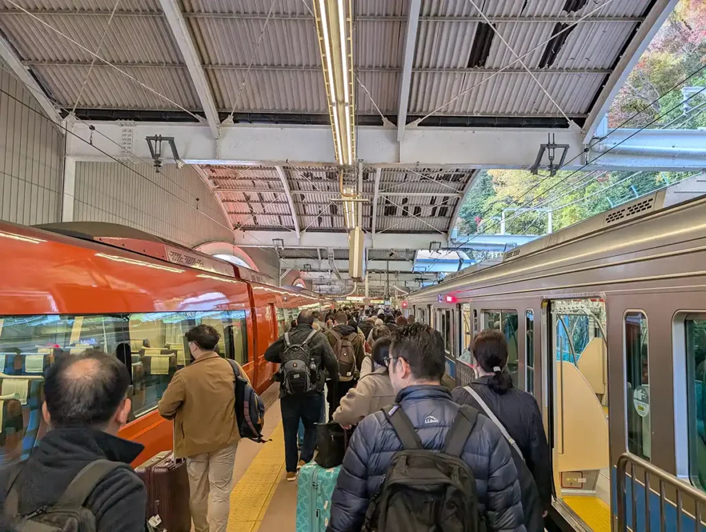 Arriving at Hakone-Yumoto station. A busy platform has a red luxury Romancecar train at one side and a silver local train on the other. 